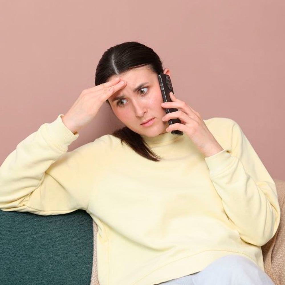 A woman in a yellow sweater sits on a couch, holding a phone to her ear with one hand and touching her forehead with the other, looking stressed or worried.