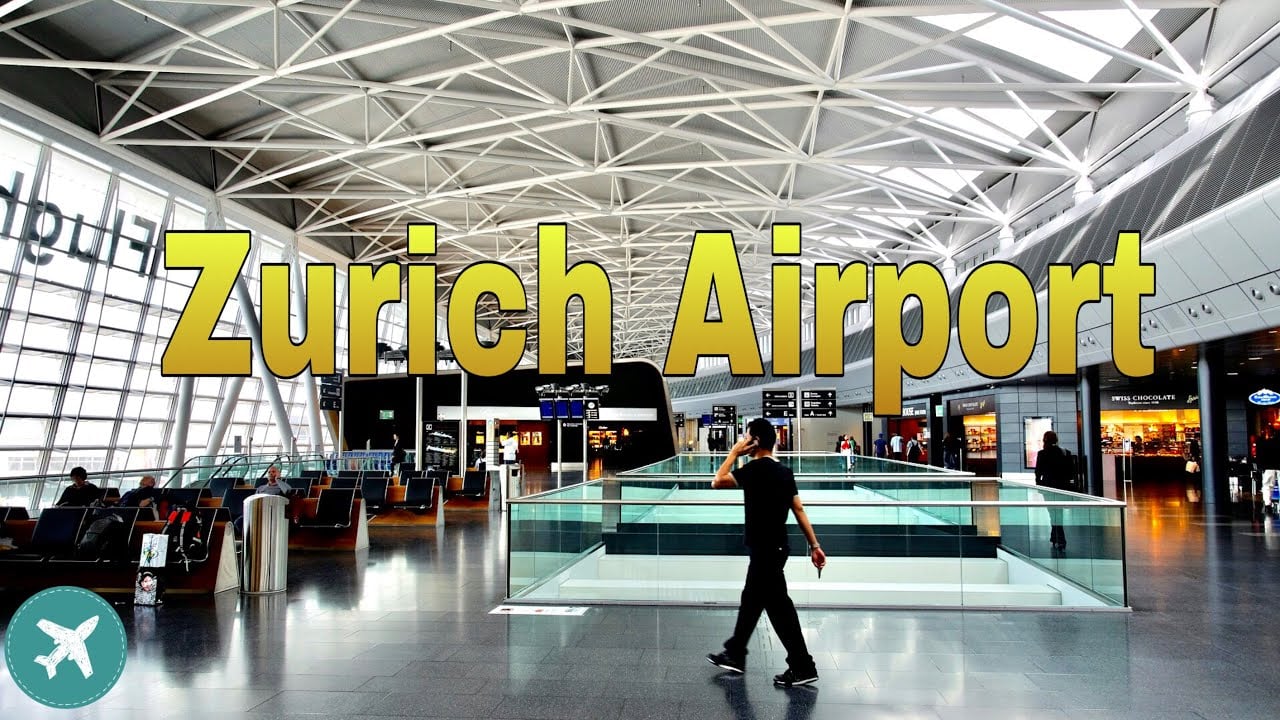 A person walks through a spacious, modern airport terminal with high ceilings and large windows. Yellow text reads "Zurich Airport" across the image. Travelers sit in waiting areas, and flight information displays are visible.