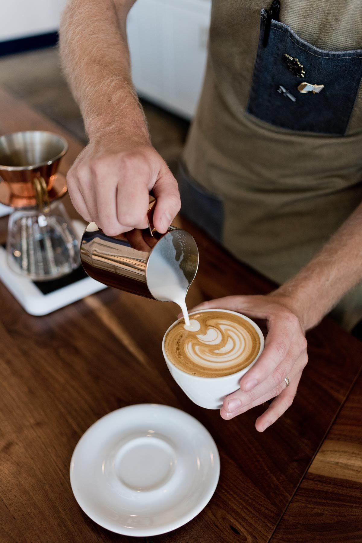 A barista pours steamed milk from a metal pitcher to create latte art in a cup of coffee on a wooden table, with an empty saucer nearby.