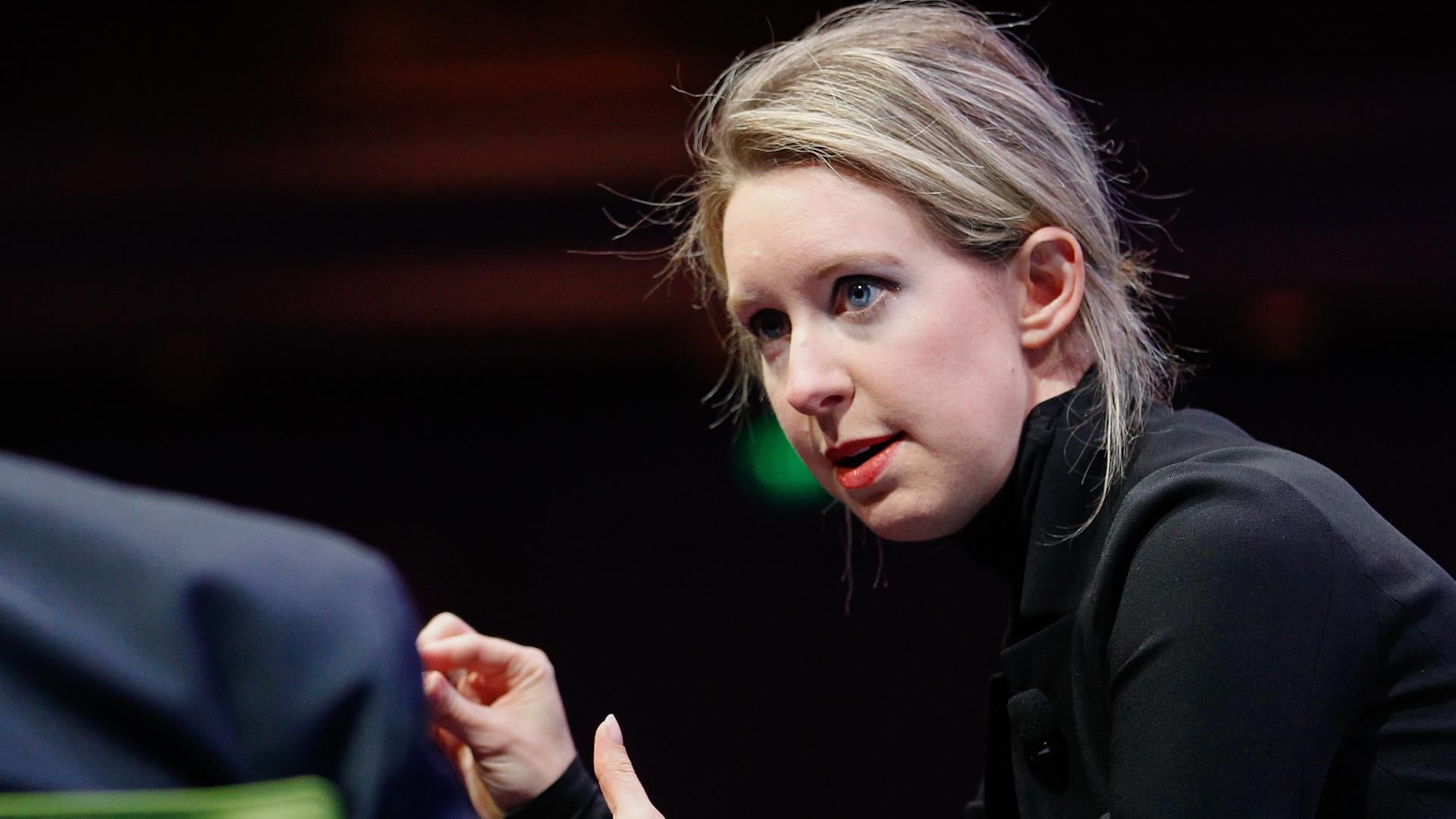 A woman with blonde hair pulled back, wearing a black turtleneck, gestures with her hand while speaking in a dimly lit setting.