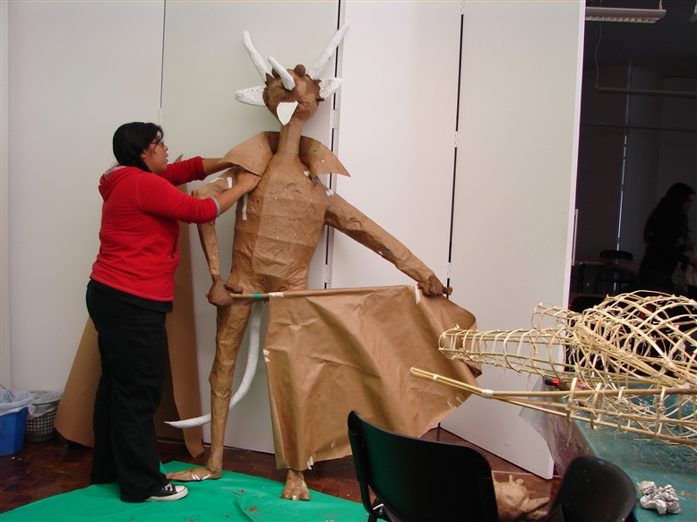 A person in a red sweater works on a large, humanoid figure made of brown paper with white horns and tail in an art studio. Art supplies and another unfinished figure are visible nearby.