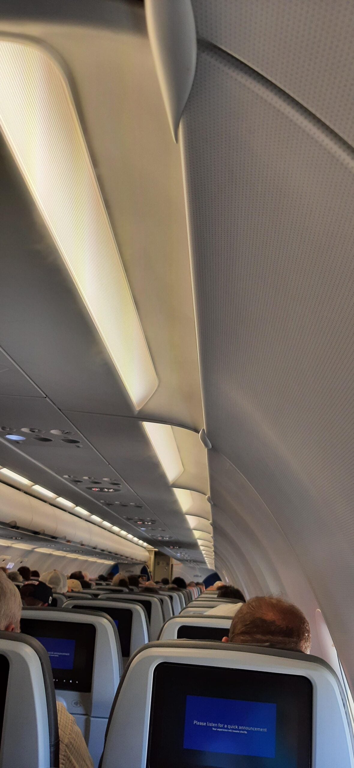 Interior of an airplane cabin with passengers seated, overhead compartments closed, and personal screens on seat backs. An illuminated sign can be seen above, and the cabin is softly lit.
