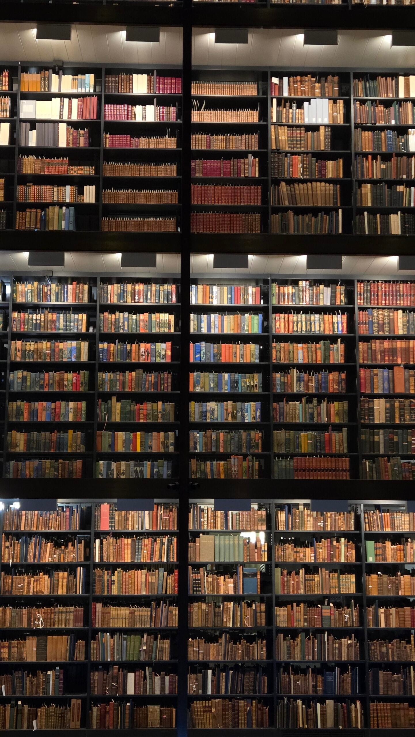 Three tall shelves filled with rows of colorful, tightly packed books reach up to the ceiling in a dimly lit library, viewed from the ground looking upward.