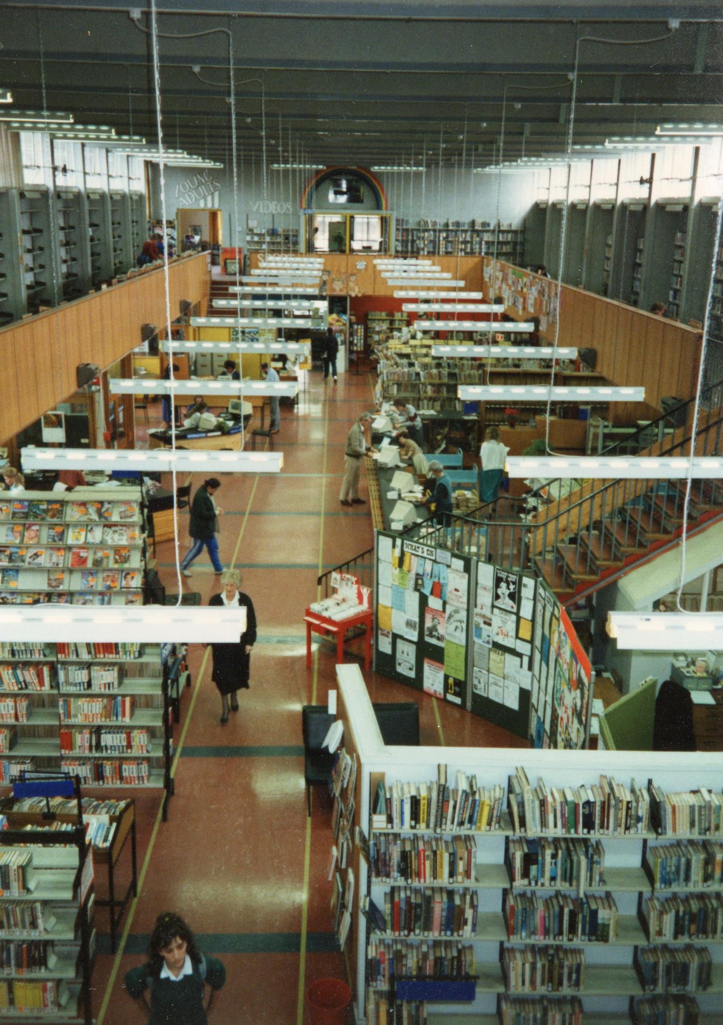 View of a spacious library interior from above, with bookshelves, reading tables, people studying, walking, and books displayed along the walls. Bulletin boards and bright overhead lights are also visible.