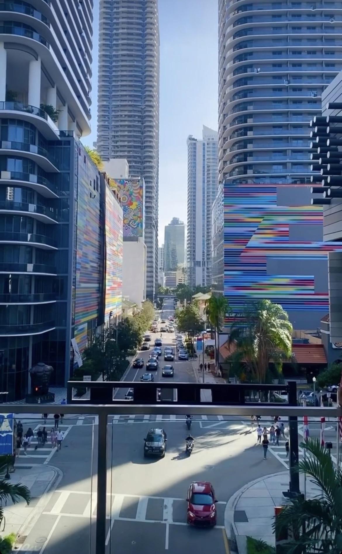 A city street scene with tall modern buildings on both sides, some featuring colorful murals, cars driving and parked, pedestrians crossing, and palm trees lining the sidewalks under a clear sky.