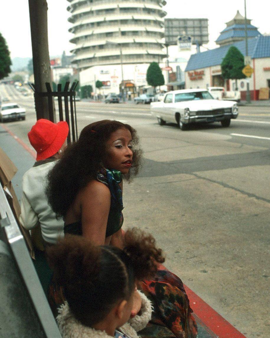 Three people sit on a sidewalk bench near a street; a woman in a red hat, another woman with long hair, and a child with a bun. Classic cars and a round building are visible in the background.