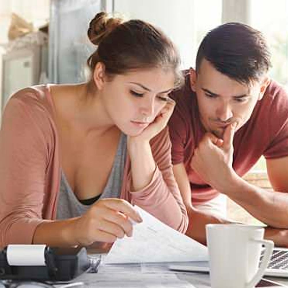 A woman and a man sit closely together at a table, looking thoughtfully at documents. The woman holds a paper, and a coffee mug and calculator are on the table in front of them.