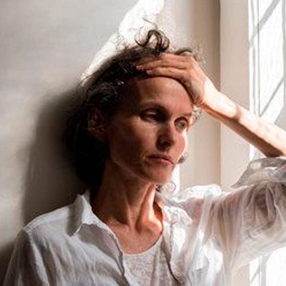 A woman in a white shirt leans against a wall, looking tired or stressed, with her hand resting on her forehead as sunlight streams in through a nearby window.