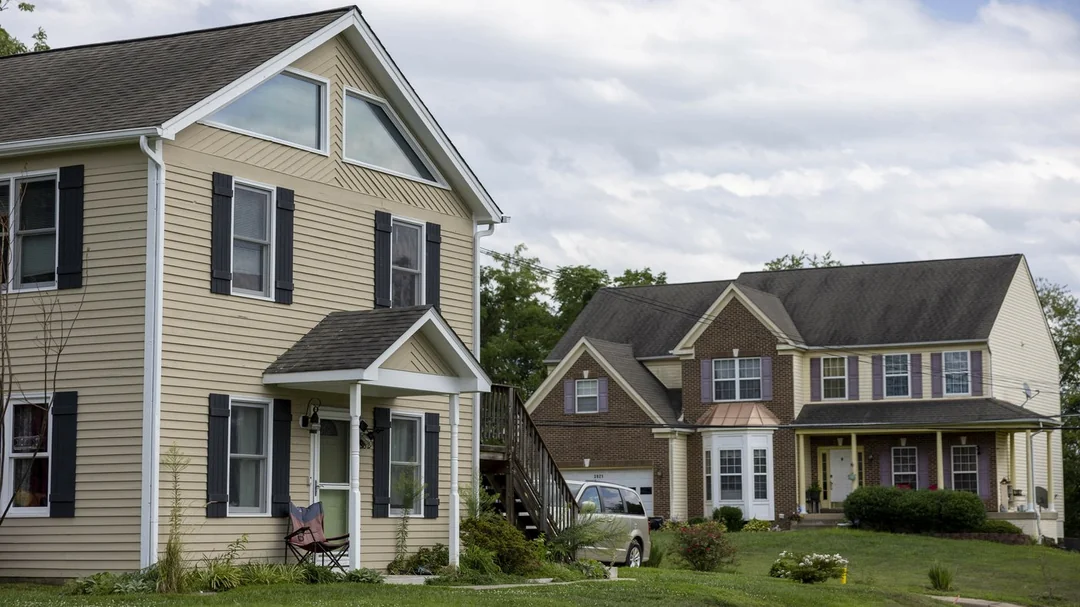 Two suburban houses sit on green lawns under a partly cloudy sky. The house on the left is beige with black shutters, while the larger house on the right is brick and tan with a porch and parked car in the driveway.