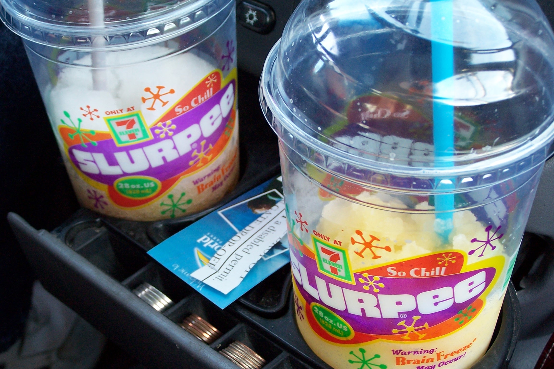 Two 7-Eleven Slurpee cups with domed lids and straws sit in a car cup holder. One drink is white, the other is orange-yellow. A card and coins are also visible in the cup holder slot between the cups.