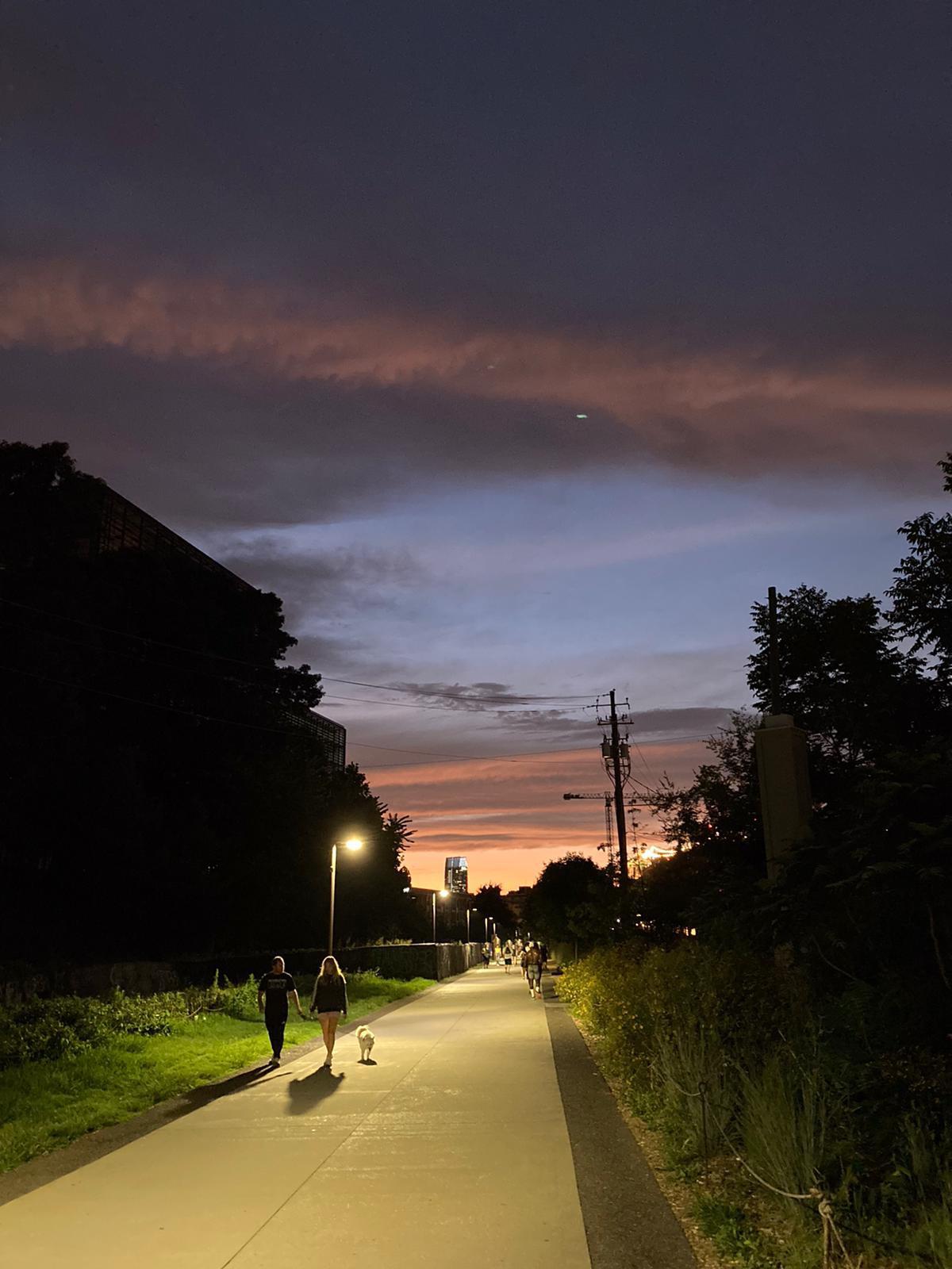A paved path at dusk with streetlights, people walking, and a small dog. The sky shows layers of dark clouds and a colorful sunset, framed by silhouettes of trees and buildings.