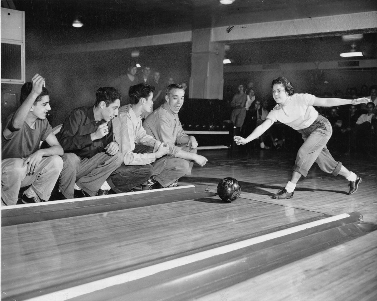 A woman bowls as four young men watch closely from the side of the lane, cheering and reacting enthusiastically. The scene takes place in a vintage bowling alley.