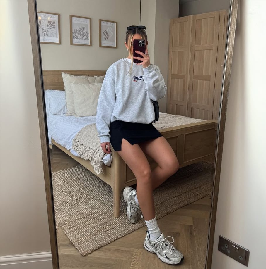 A person takes a mirror selfie in a bedroom, wearing a gray sweatshirt, black mini skirt, white socks, and sneakers. The room has a bed with beige bedding, wooden furniture, and framed art on the wall.