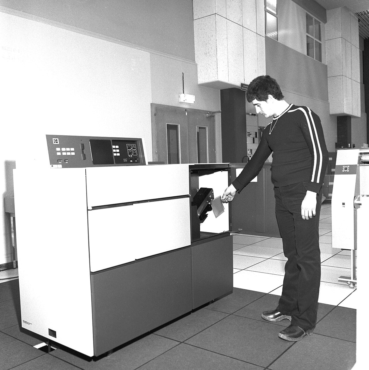 A man in dark clothing operates a large vintage computer printer, removing a stack of paper in a retro office setting with tiled floors and plain walls.