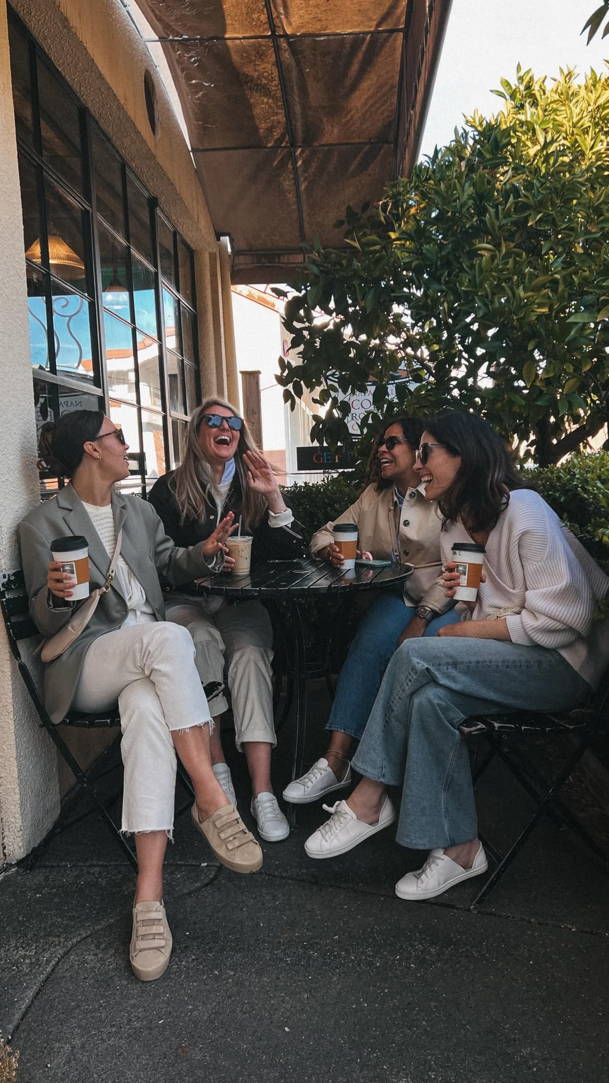 Four women sit outside at a café table, laughing and enjoying coffee together. They are dressed casually and appear to be having a lively, friendly conversation on a sunny day.