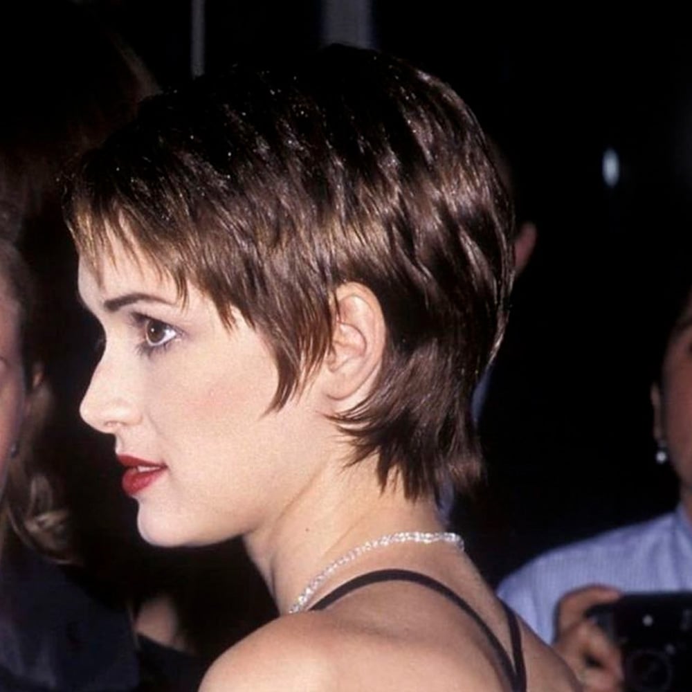A woman with short, layered brown hair and red lipstick is shown in profile at a formal event, wearing a black spaghetti strap top. The background is dark and blurred, with other people partially visible.