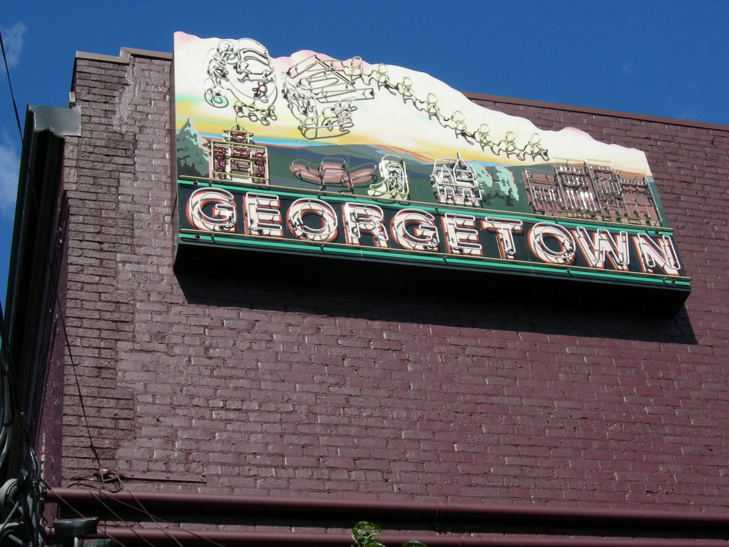 A neon sign reading "GEORGETOWN" with illustrations of buildings and faces is mounted on a dark brick wall against a clear blue sky.