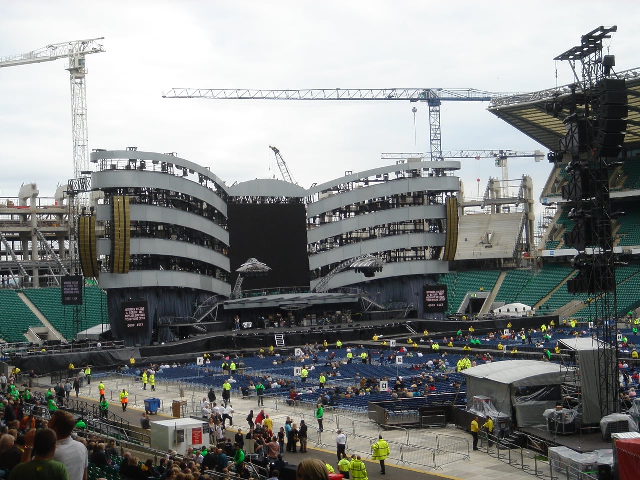 A large outdoor concert stage with curved silver structures, a central black screen, lighting rigs, and cranes in the background. Security staff and people are gathered in front of the stage in a stadium.