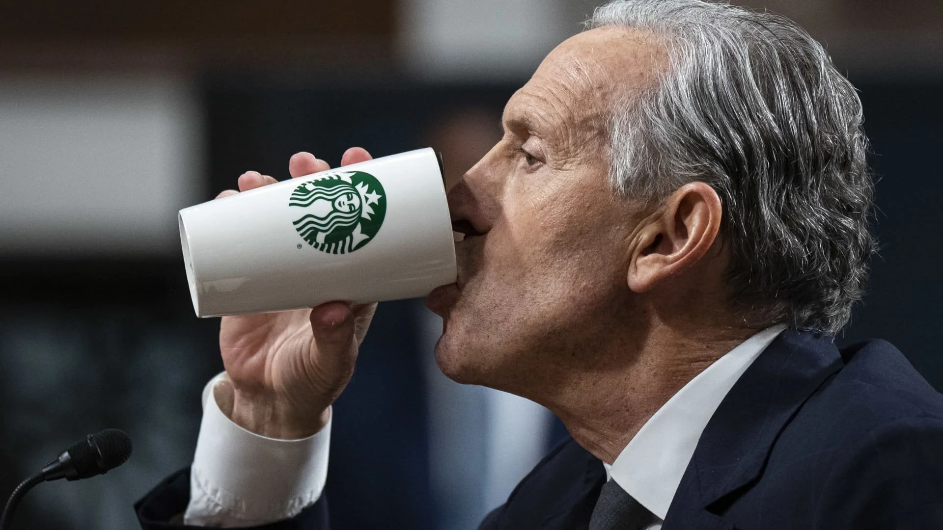A man in a suit drinks from a white Starbucks cup, holding it near his mouth, with a microphone visible in the foreground.