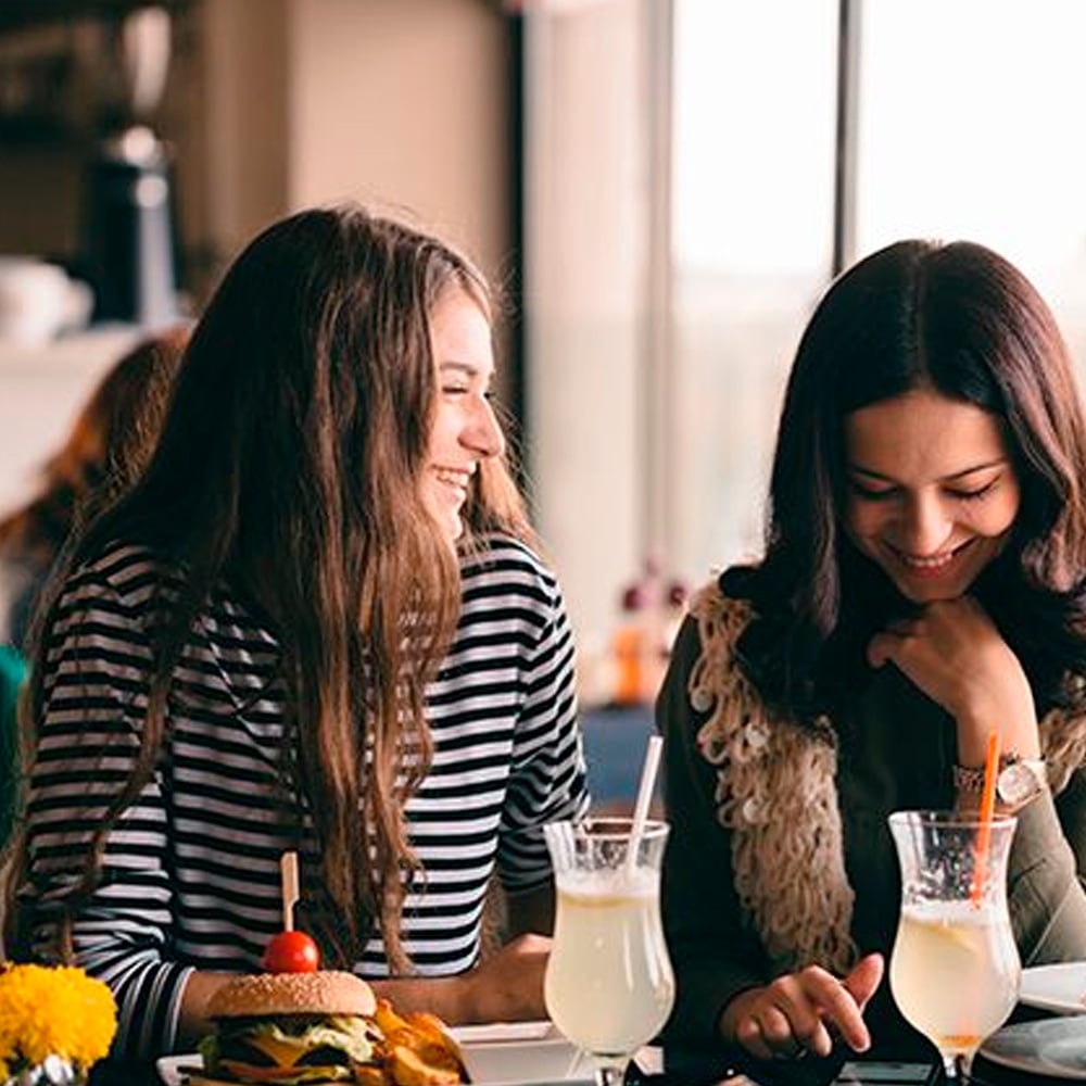 Two young women sit at a café table, smiling and enjoying drinks with lemon slices. One has a burger and fries in front of her. They appear to be having a friendly conversation in a bright, casual setting.