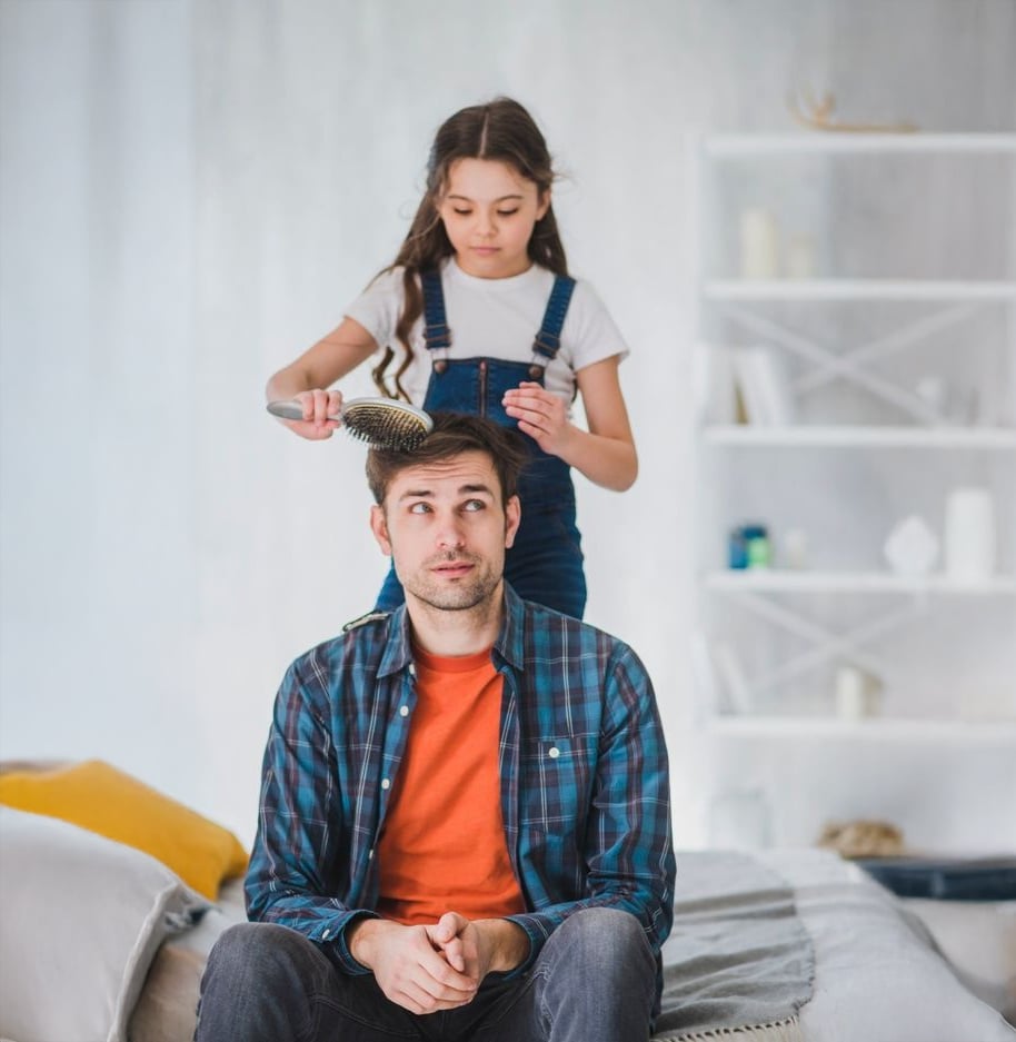 A young girl brushes the hair of a man sitting on a bed. The man looks upward with a playful expression while the girl focuses on brushing. They are in a bright, cozy room with shelves and a bed in the background.