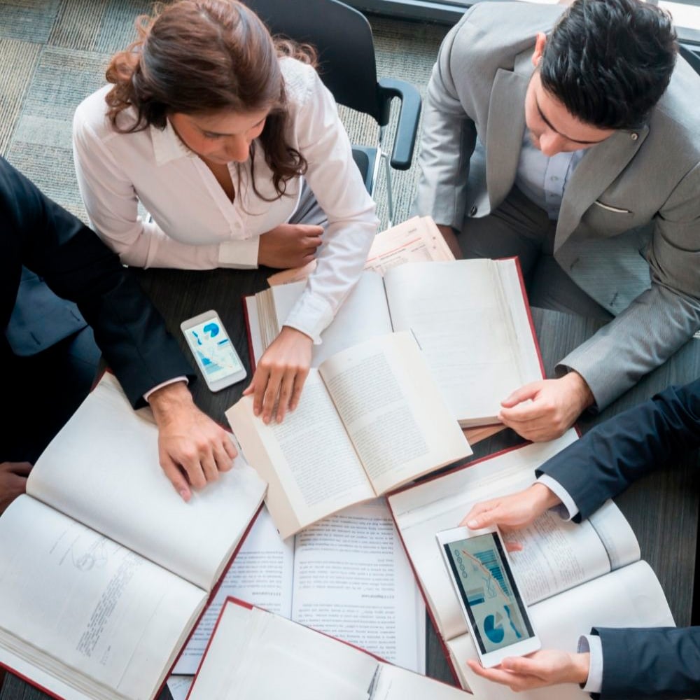 Four people in business attire sit around a table with open books, documents, a phone, and a tablet displaying charts, collaborating on work or study.