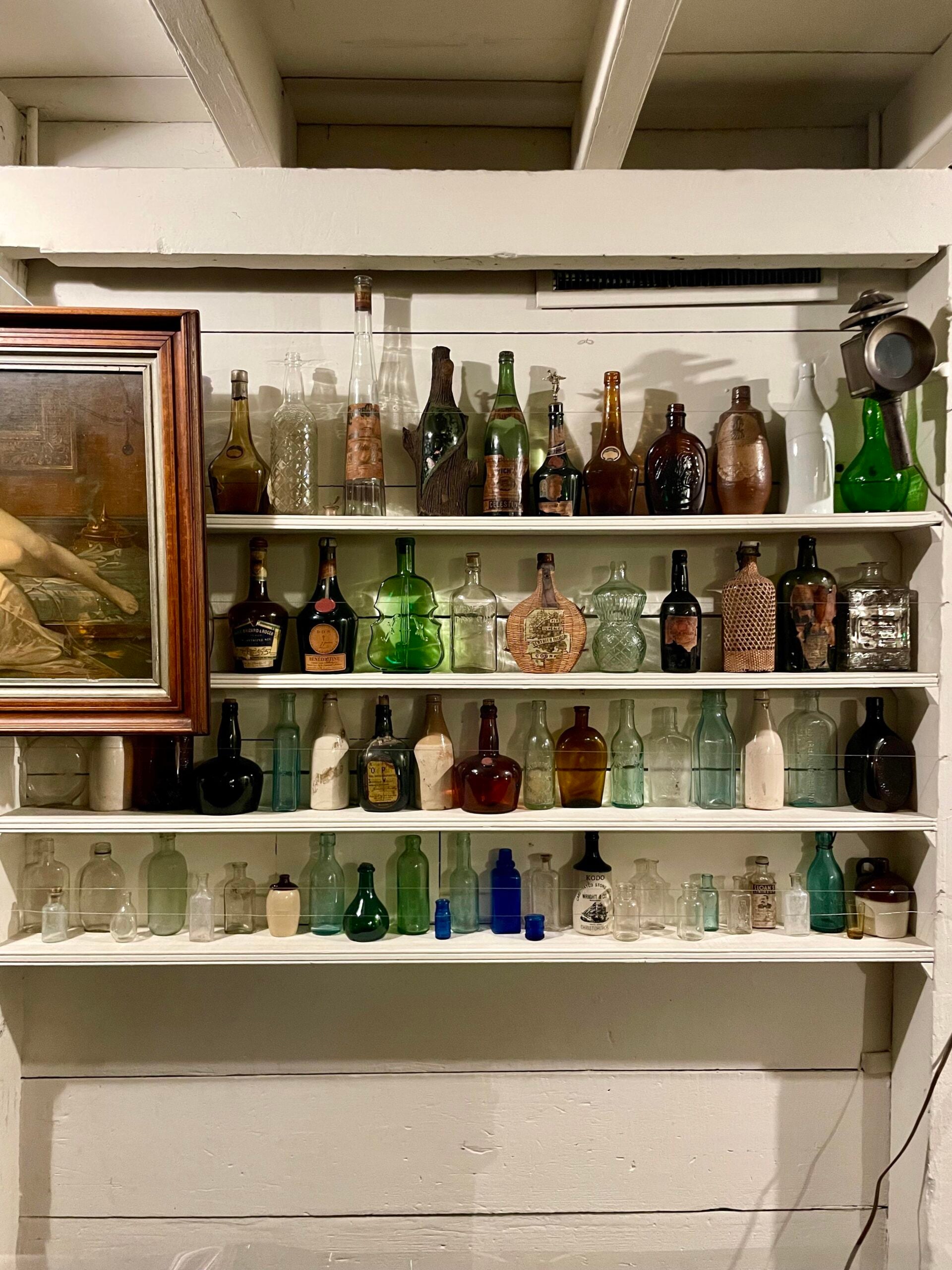 Shelves filled with a variety of vintage glass bottles and jars in different shapes, sizes, and colors, some with labels. A framed painting and an old camera are displayed on the left and upper right.
