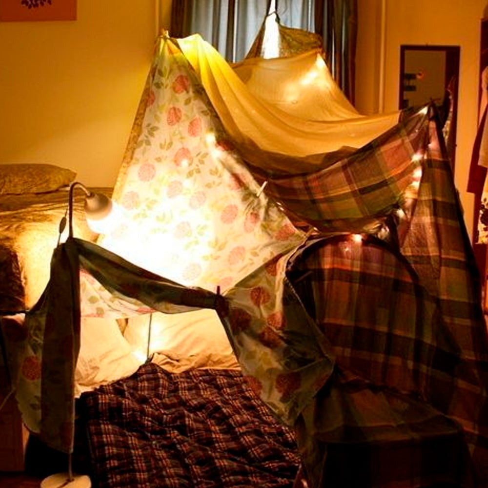 A cozy indoor blanket fort is built with patterned sheets and lit by string lights, set up next to a bed in a warmly lit bedroom. Blankets and pillows are spread inside the fort for comfort.