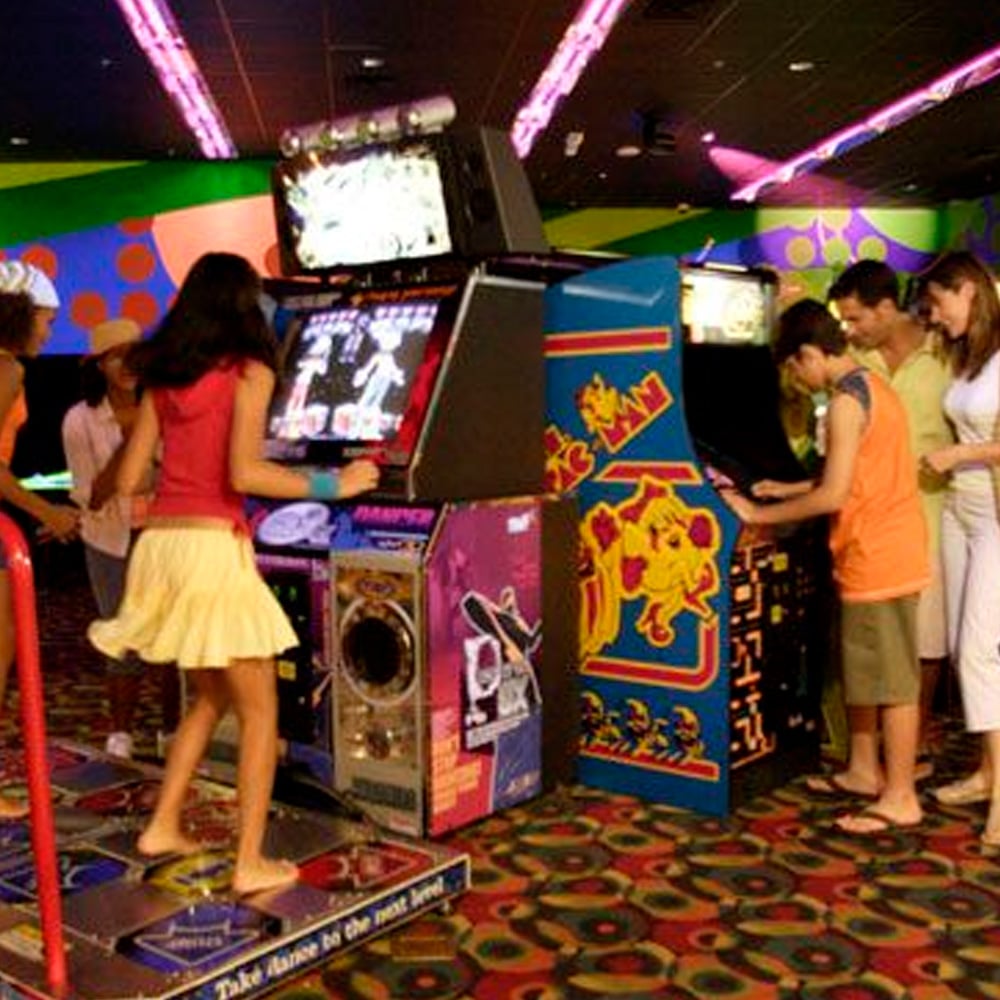People play arcade games in a colorful, neon-lit arcade. A girl uses a dance machine, while others play classic games like Pac-Man. The floor is covered with patterned carpet.
