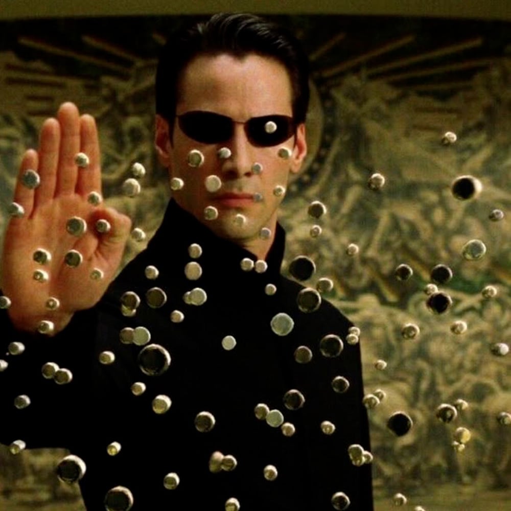 A man in black sunglasses and a black outfit holds up his hand, stopping multiple bullets mid-air in front of him, with a blurred mural in the background.