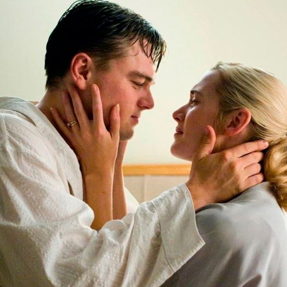 A man and a woman stand close together, gently holding each other's faces and gazing into each other's eyes with affection, both wearing light-colored clothing.