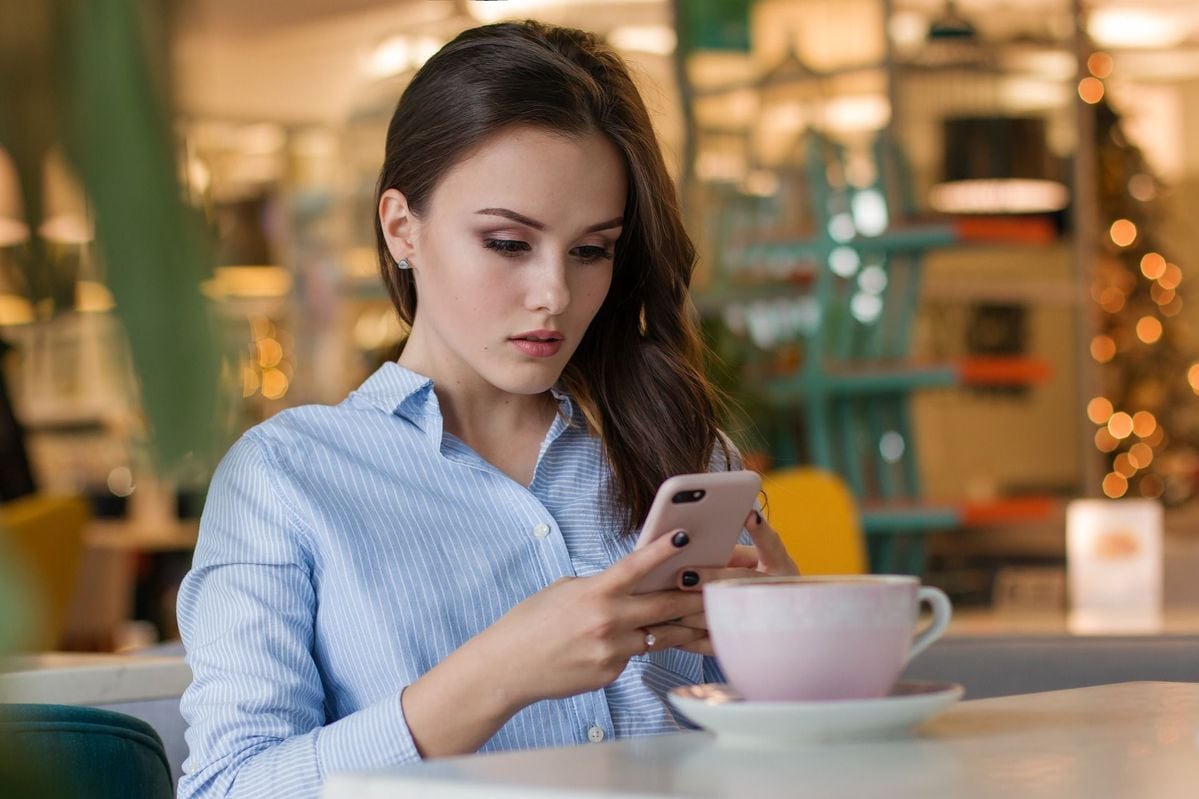 A young woman with long brown hair sits at a cafe table, wearing a light blue striped shirt, looking intently at her smartphone. A large pink coffee cup is in front of her, and the background is softly blurred.