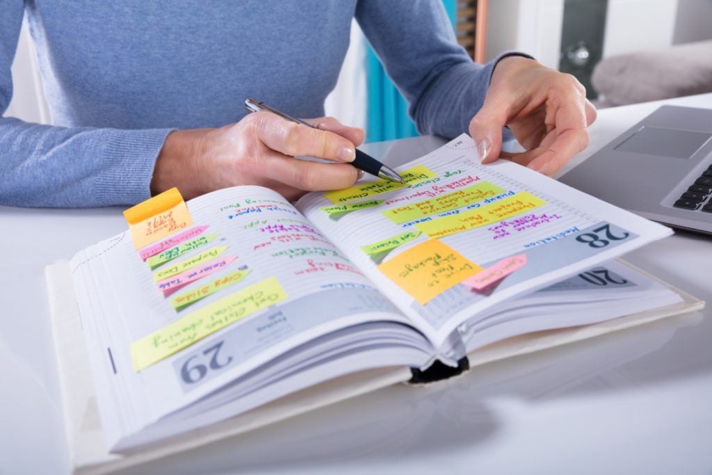 A person writes in a planner filled with colorful sticky notes and handwritten tasks, sitting at a white desk next to a laptop. The scene suggests organization and planning.