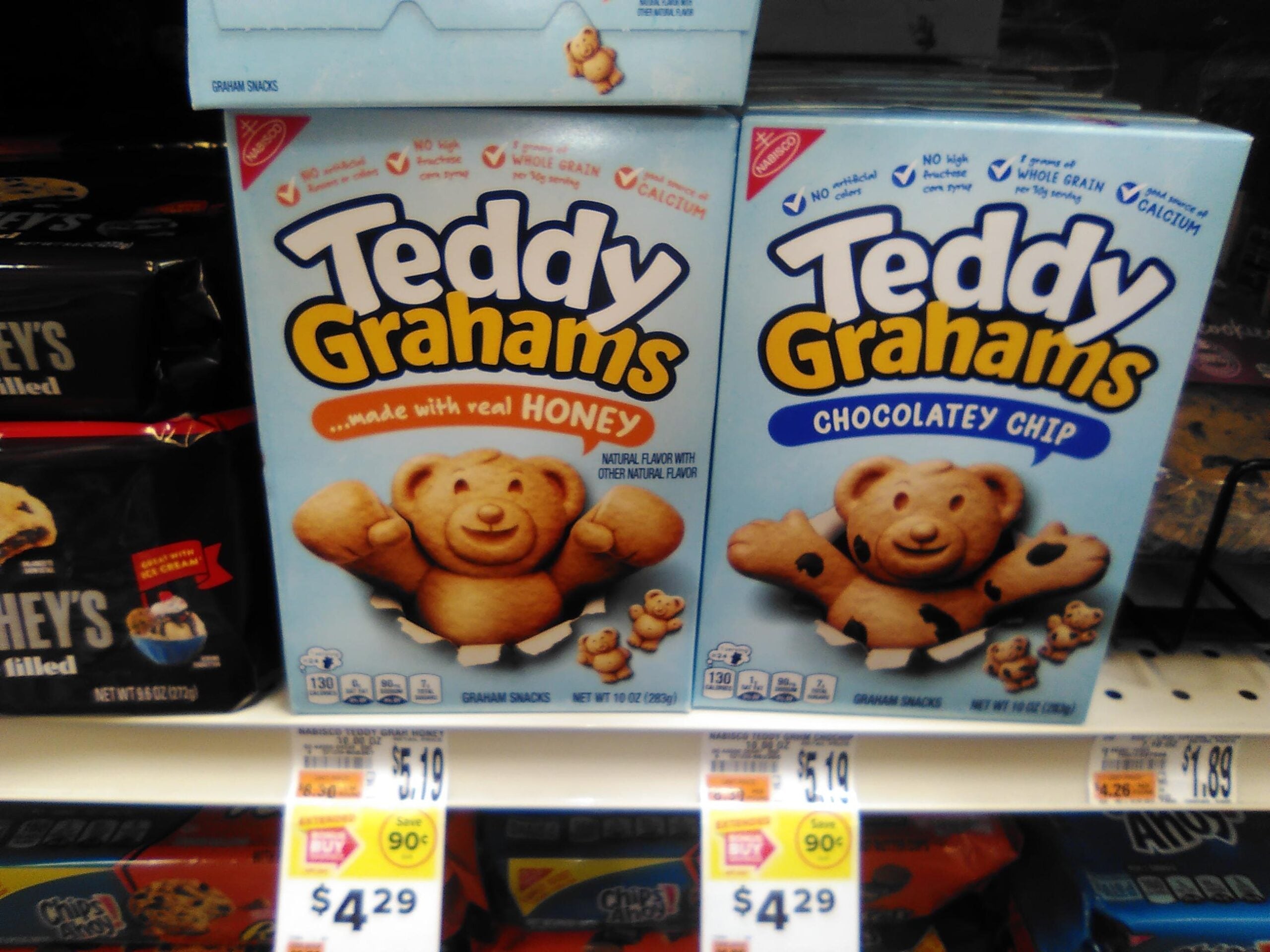 Two boxes of Teddy Grahams snacks, Honey and Chocolate Chip flavors, are displayed on a grocery store shelf with price tags below them. Each box features a smiling bear-shaped graham cracker on the front.