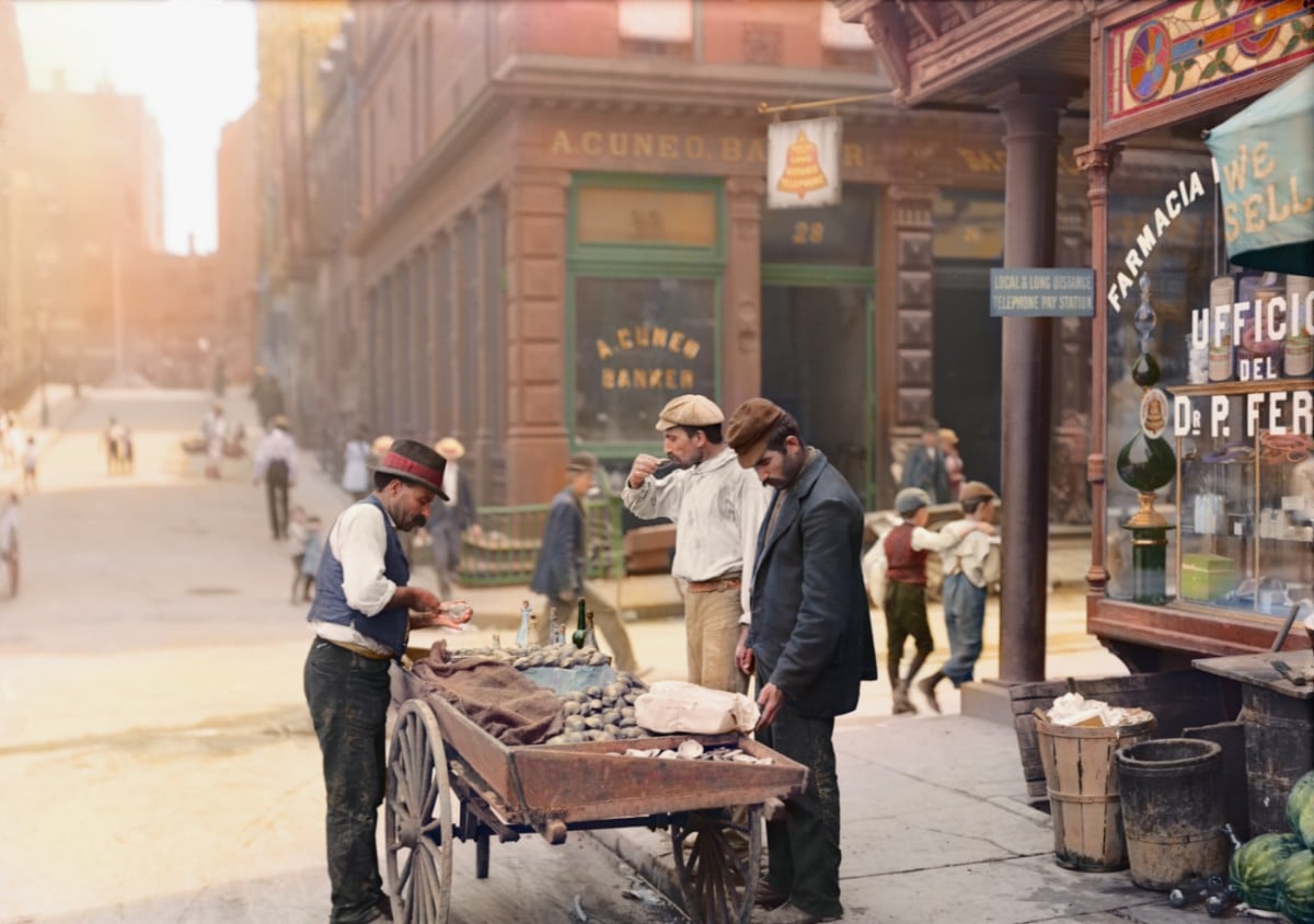 A street vendor in early 1900s attire sells goods from a cart on a busy city street, surrounded by men and children, with historic storefronts and buildings in the background.