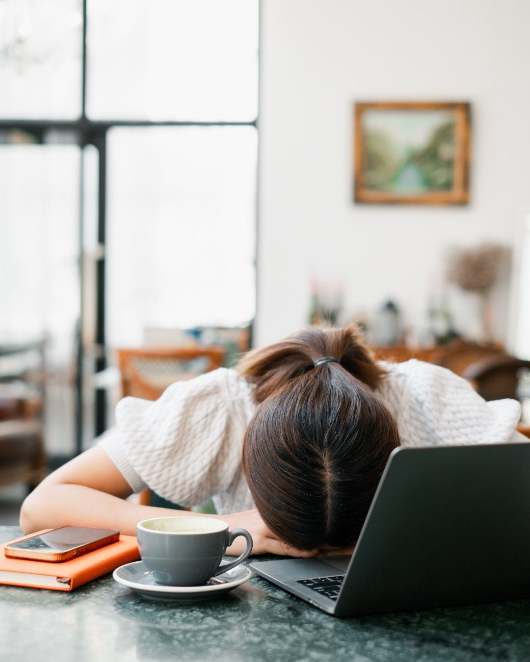 A person with brown hair rests their head on a table in front of an open laptop, looking tired or overwhelmed. A cup of coffee and a smartphone sit nearby in a bright, cozy room.