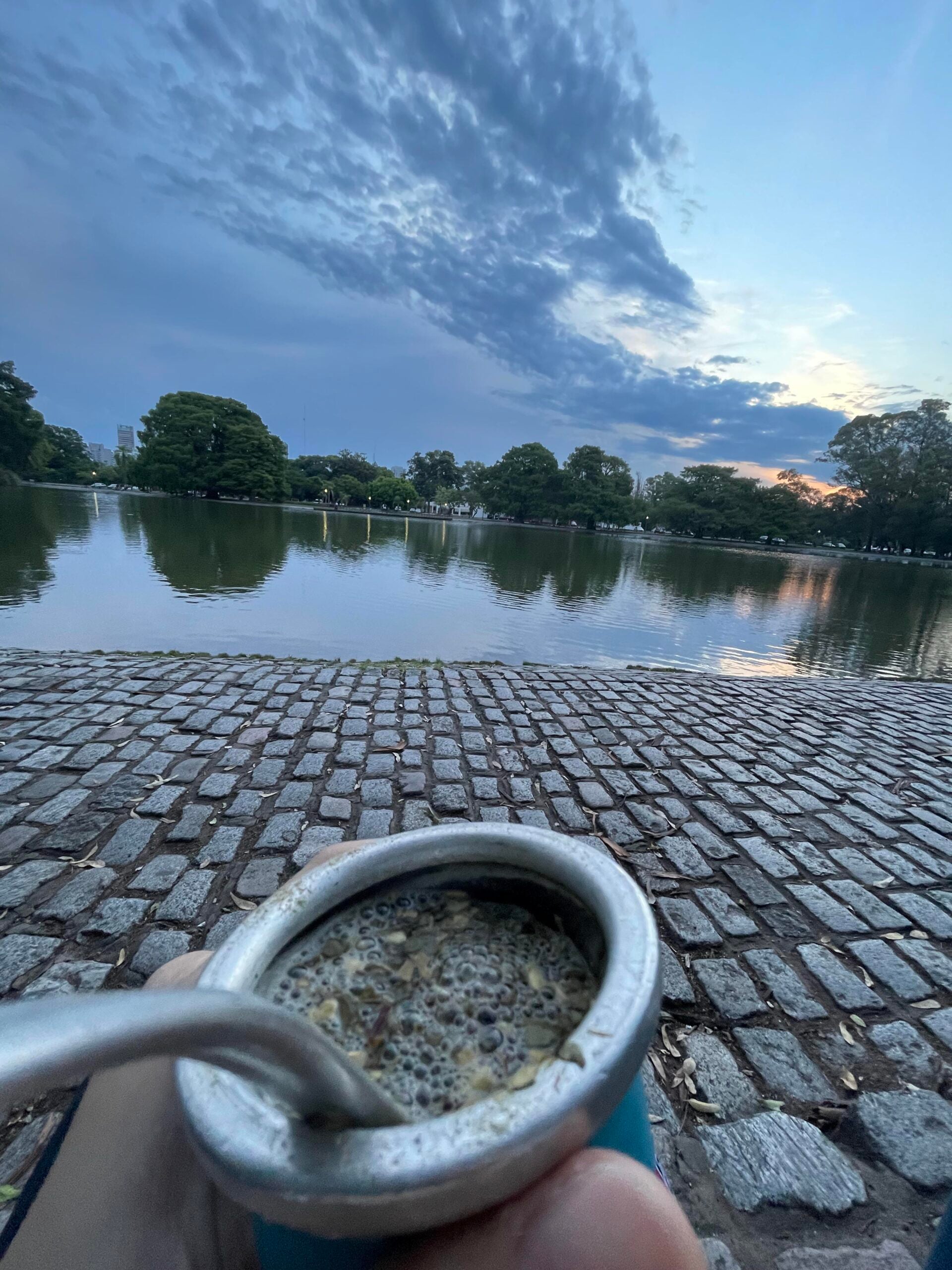 A person holds a mate gourd with a metal straw by a cobblestone path next to a calm lake at sunset, with trees and a cloudy sky in the background.