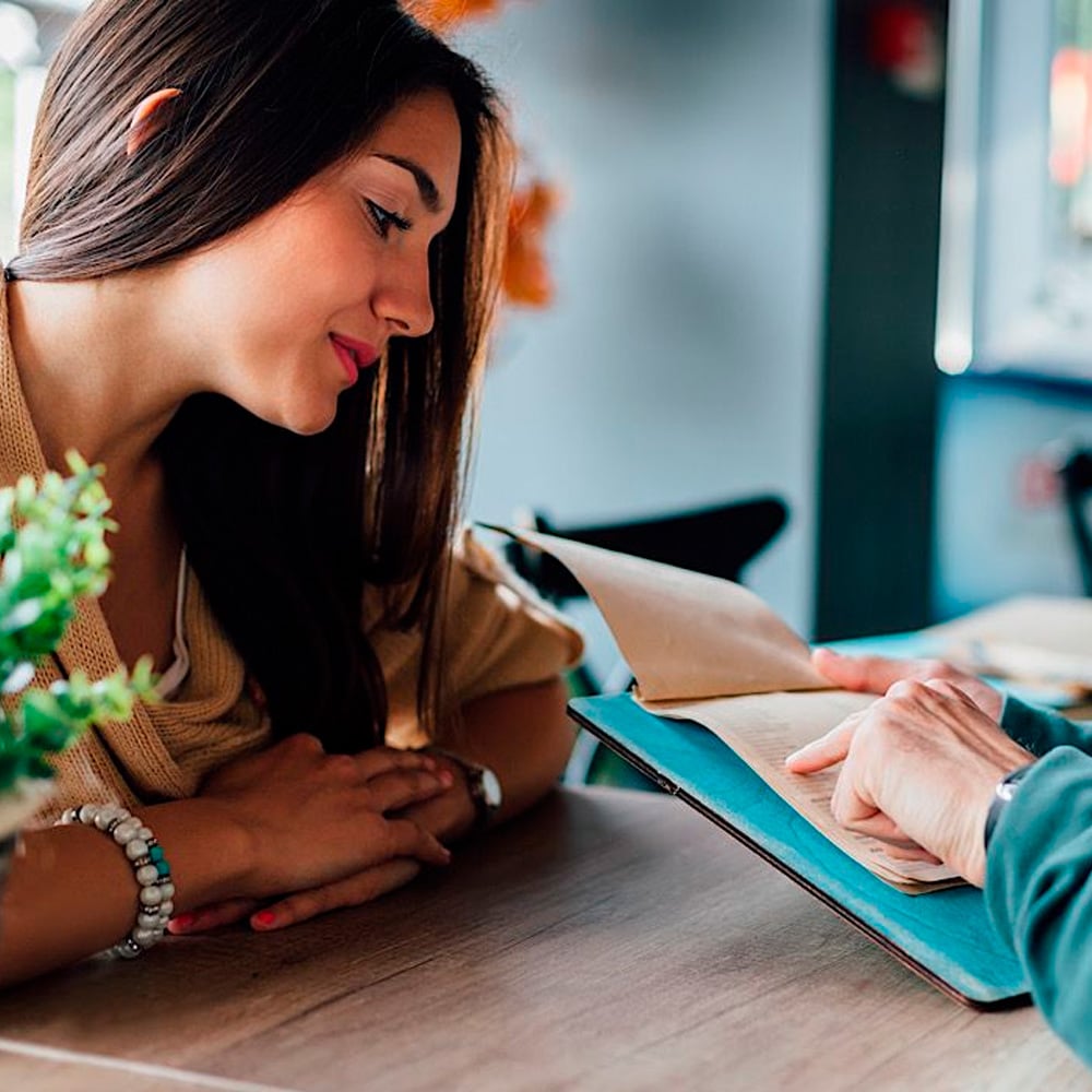 A woman with long brown hair sits at a table in a cafe, looking at a menu held by another person. She appears to be considering options, with her hands resting on the table and a small plant in the foreground.