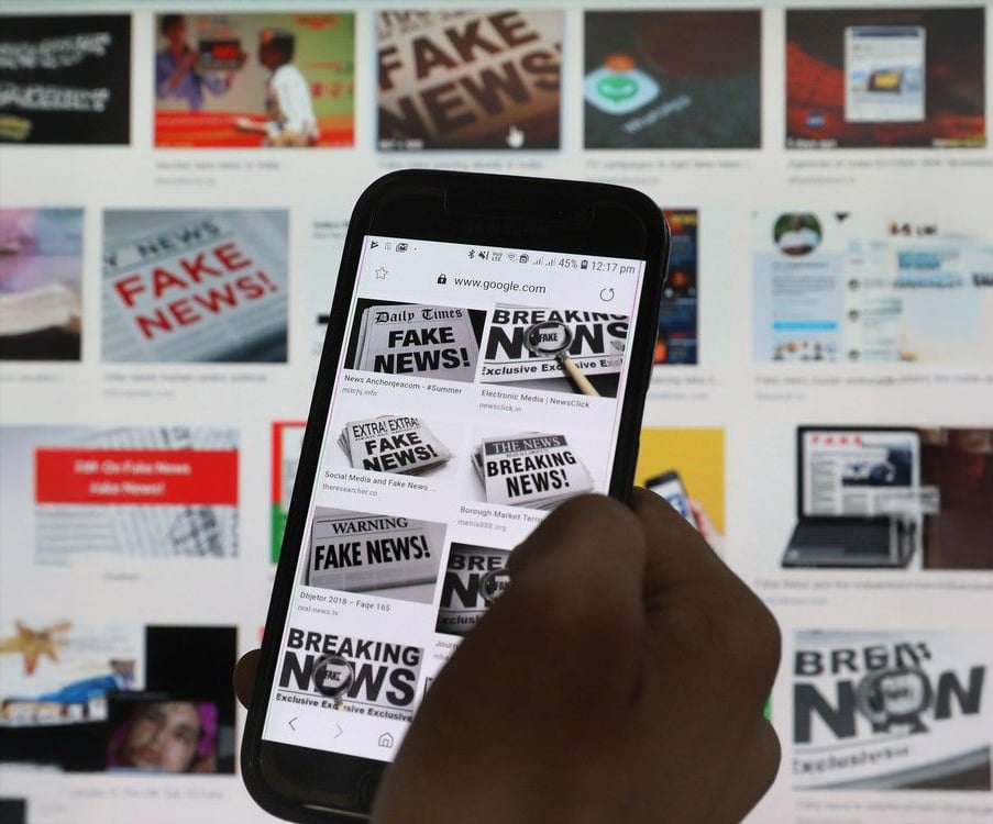 A person holds a smartphone displaying news headlines with phrases like "FAKE NEWS" and "BREAKING NEWS" in front of a computer screen showing similar images about fake news.