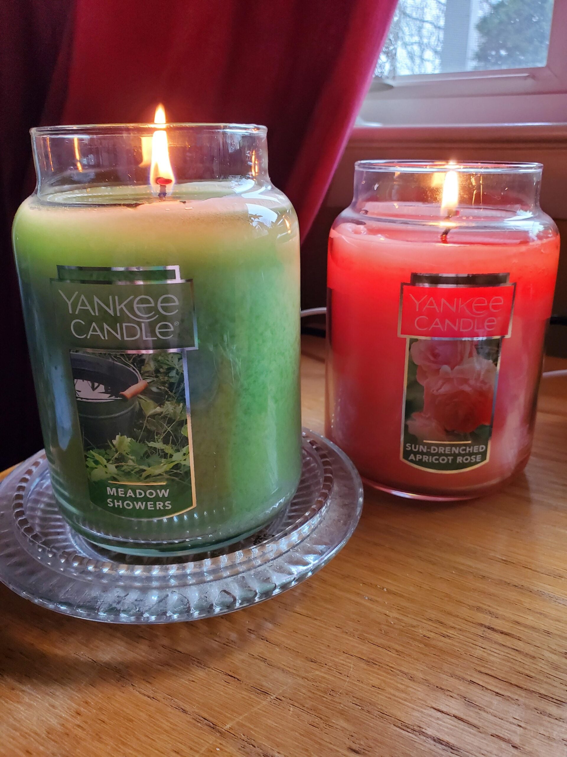 Two lit Yankee Candle jars sit on a wooden surface by a window. The green candle is labeled "Meadow Showers," and the red candle is labeled "Sun-Drenched Apricot Rose." A red curtain hangs in the background.