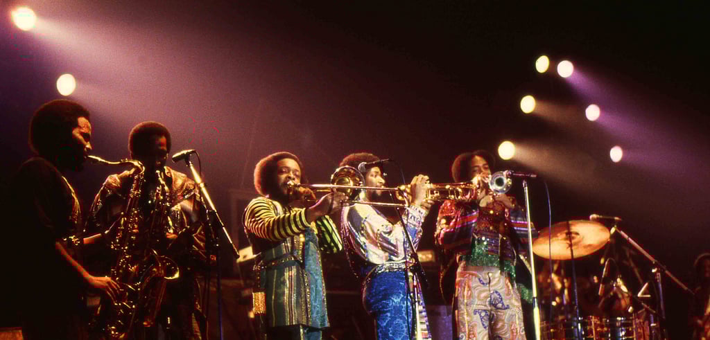A band performs on stage under bright lights, with musicians playing saxophones, trumpets, and other brass instruments, all dressed in colorful, retro outfits. Drums and additional band members are visible in the background.