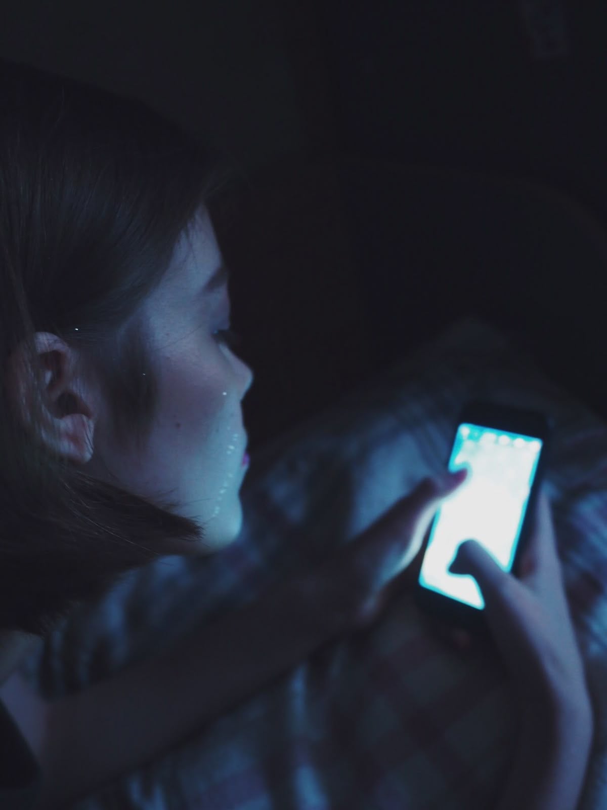 A person lies on a bed in a dimly lit room, illuminated by the light of a smartphone they are holding and looking at closely. Their face is partially visible in the glow of the screen.
