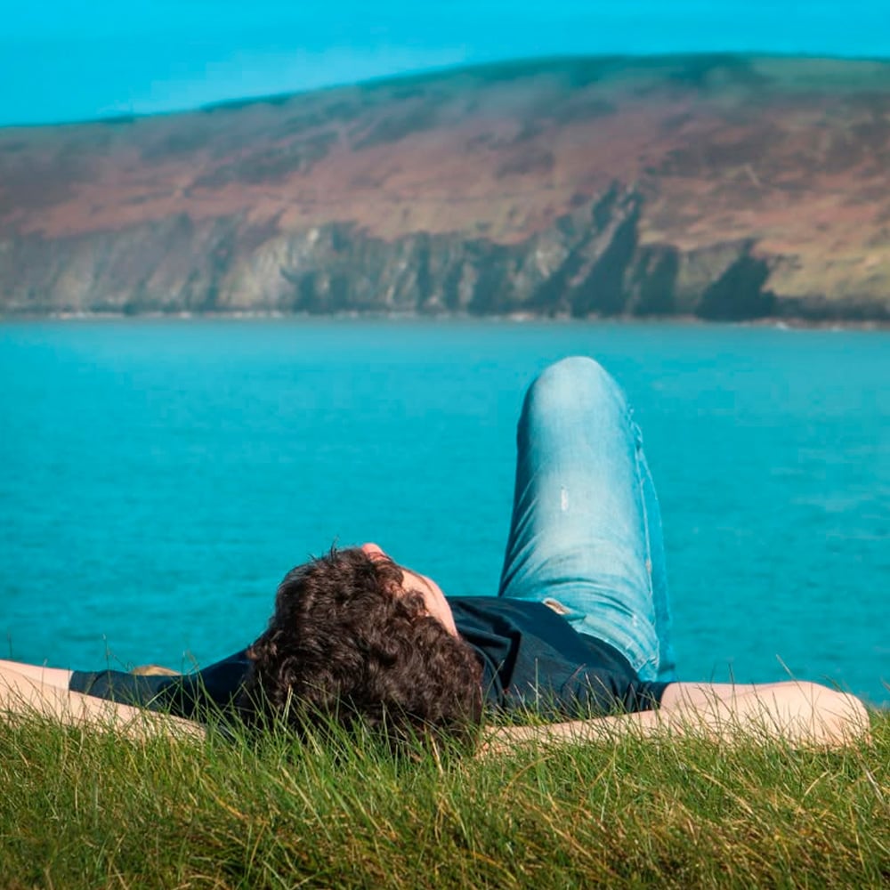 A person lies on green grass facing a bright blue lake, with hills and cliffs in the background under a clear sky.