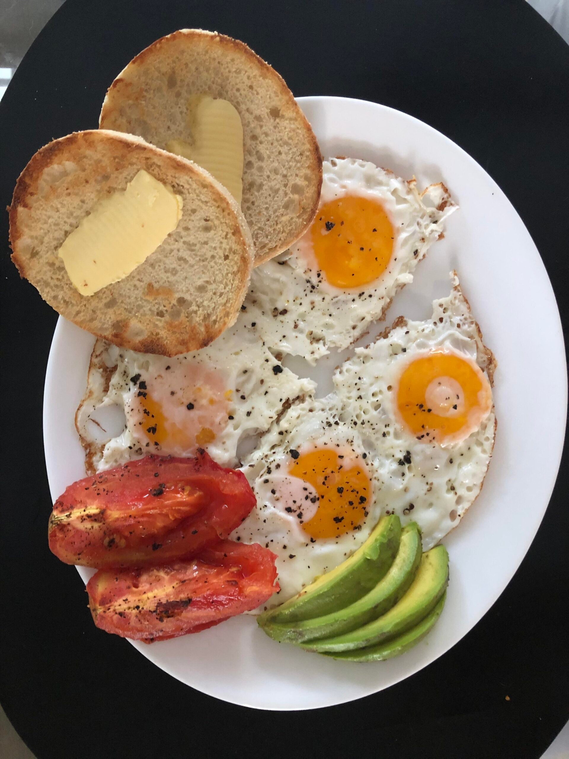 A white plate with three sunny-side-up eggs, sliced avocado, sliced tomato, and an English muffin with butter on a black surface.