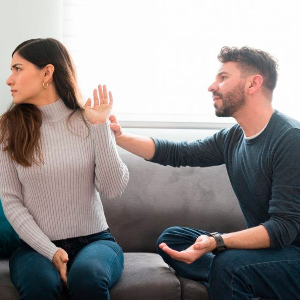 A woman sits on a couch, turning away and holding up her hand as if to refuse or stop a man next to her, who is reaching out and appears to be pleading or explaining something.