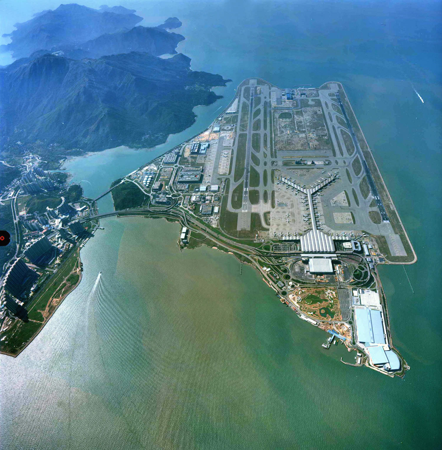 Aerial view of a large airport built on reclaimed land by the sea, with multiple runways, taxiways, terminals, and surrounding mountains in the background.
