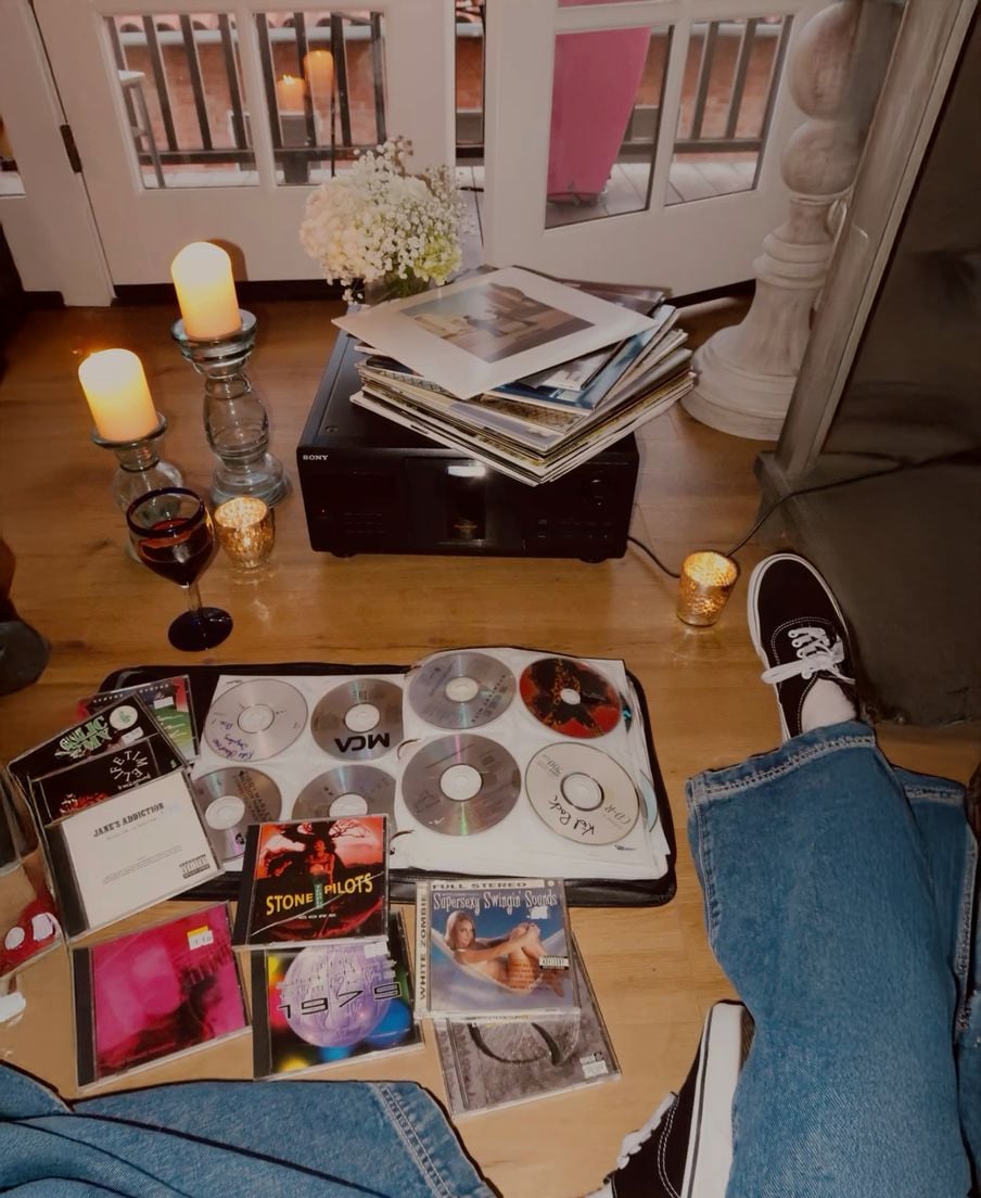 A cozy indoor scene with lit candles, a stack of vinyl records on a stereo, scattered CDs, album covers, a glass of red wine, and someone’s legs in blue jeans and black sneakers on a wooden floor.
