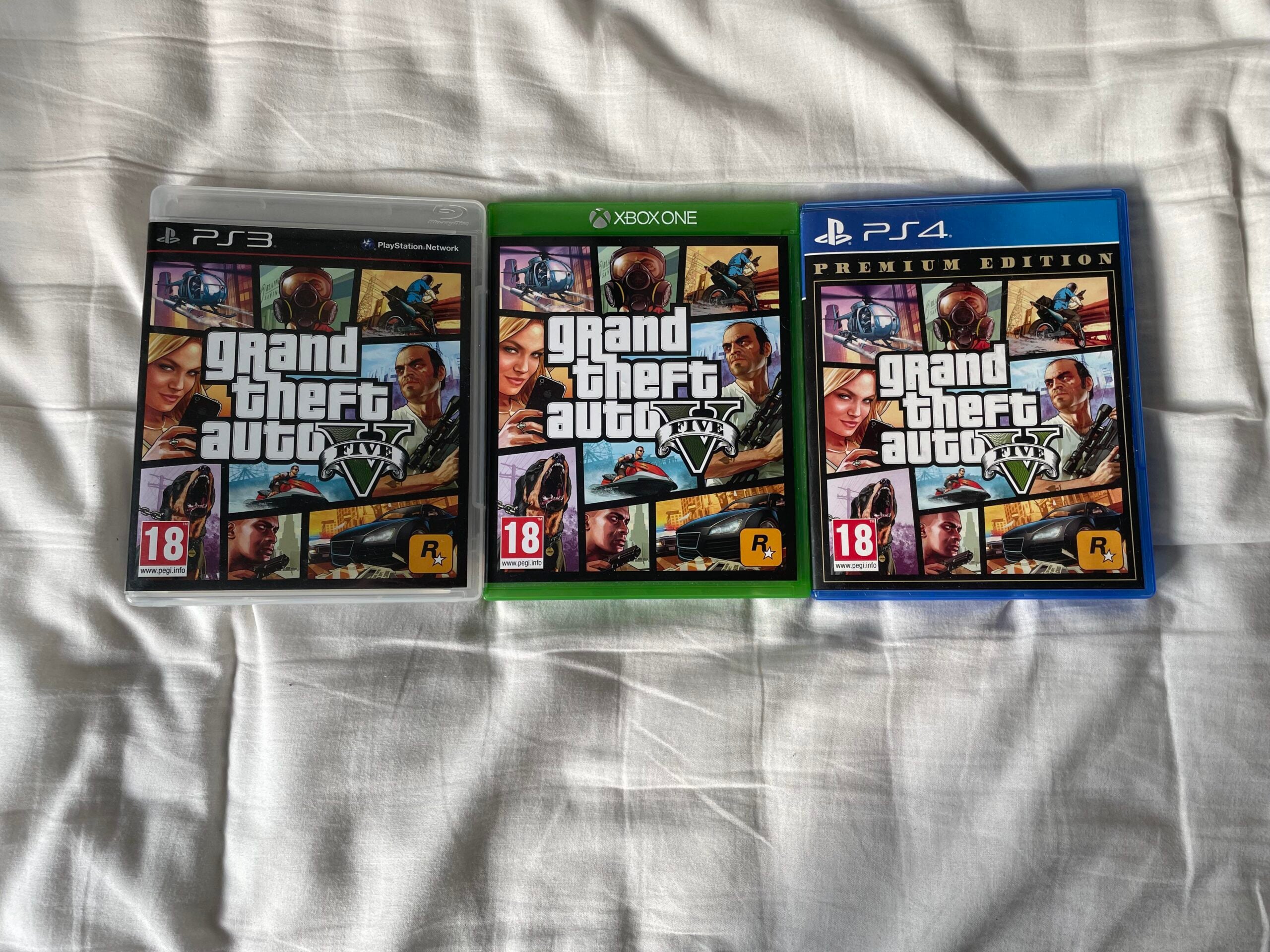 Three copies of Grand Theft Auto V are displayed on a white blanket: one for PS3 (left), one for Xbox One (center), and one for PS4 Premium Edition (right), each showing the distinctive cover art.