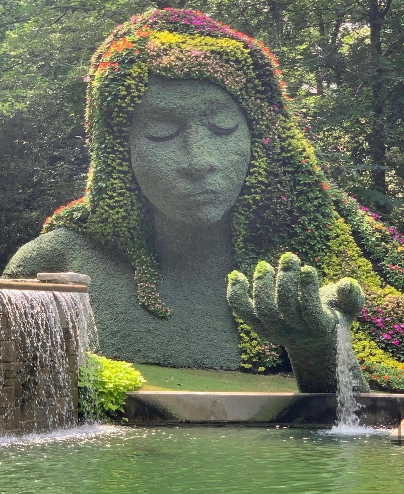A large garden sculpture of a serene woman's face and hand, covered in green plants and colorful flowers, pours water into a pond.
