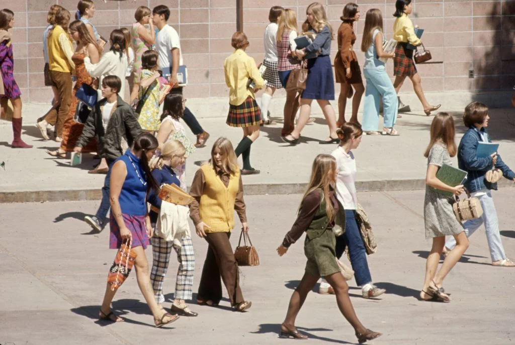 A large group of young people, mostly teenagers, walk outside on a sunny day. They wear colorful 1960s or 1970s clothing, including dresses, skirts, pants, and vests, and many carry books or bags.